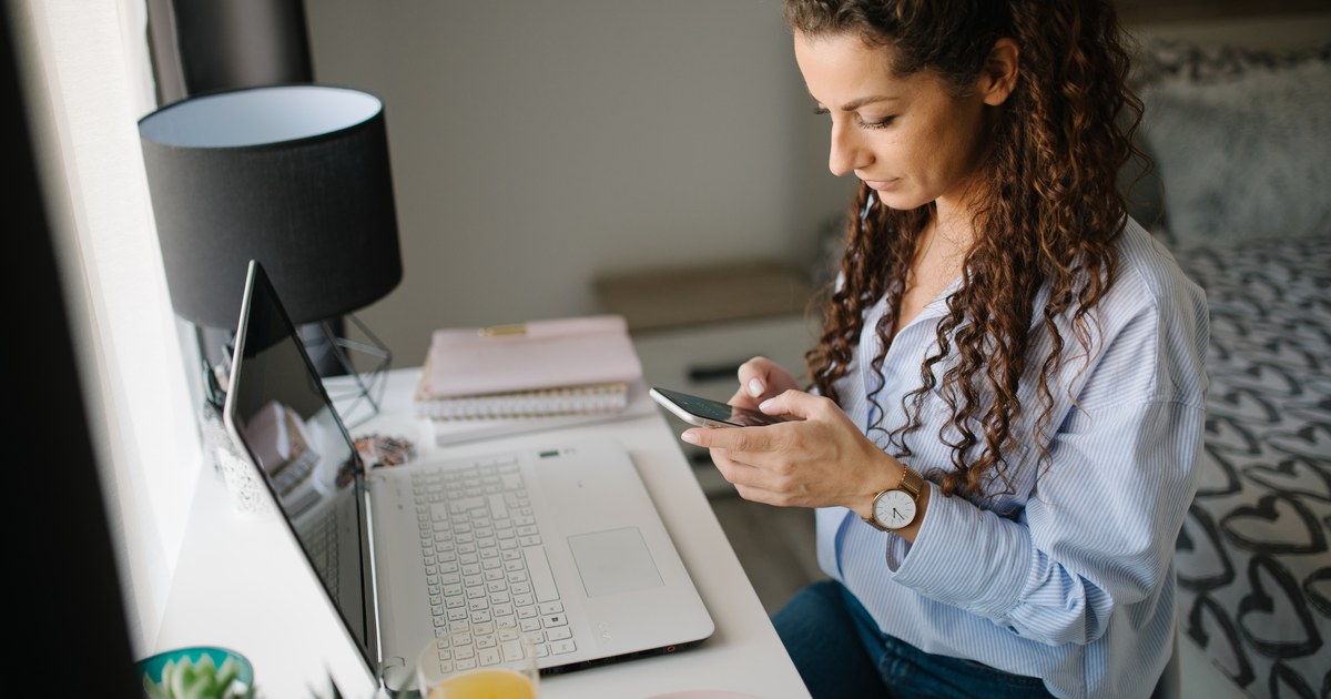 Person managing an online account on a smartphone next to a laptop at a desk