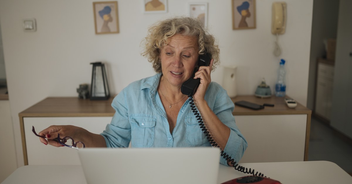 Woman at a home desk on the phone while using a laptop, reviewing her account.
