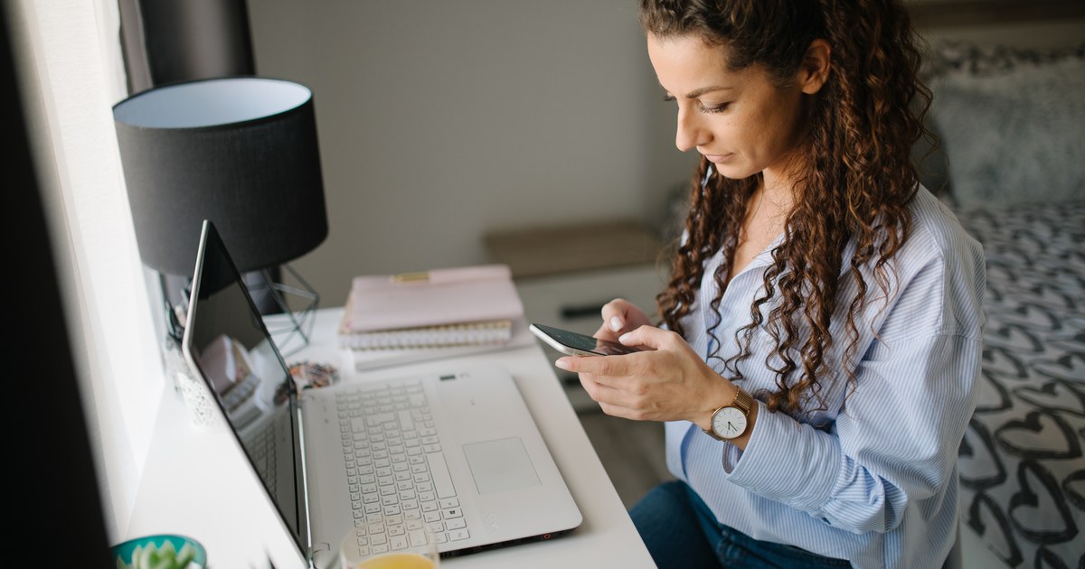 A person using a smartphone next to a laptop at a home desk, illustrating online account checks and responsible gambling tools.