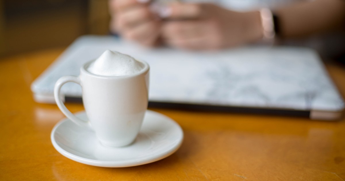A coffee cup on a wooden table in front of a person using a phone beside a laptop